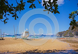 Traditional philippine boat called bangka on the beach with tree leaves on forefround. Fishing boat on seacoast in Asia.