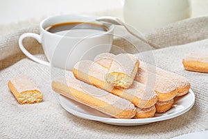 Traditional savoiardi biscuits or ladyfingers cookies on a white plate and a white cup of coffee