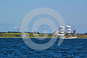 Traditional square-rigged tall ship and modern two-masted yacht at sea in front of a green seashore with fields and wind power sta