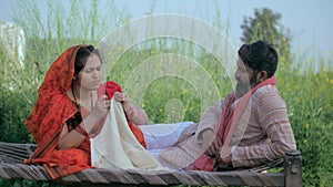 Traditional wife sewing clothes sitting on a cot while chatting with her husband - Indian farmer
