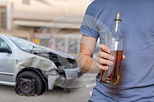 Traffic accident and alcohol concept. Drunk driver is holding bottle with alcohol in hand. Crashed car in background.