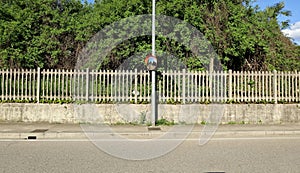 Traffic convex mirror on metal pole. Old concrete railway fence and vegetation on behind, concrete sidewalk and street in front