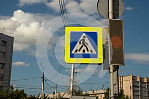 Traffic sign Pedestrian crossing hangs on a pole next to a traffic light on the background of the sky and city buildings on a