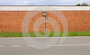 Traffic wide angle security convex mirror on a pole with brick wall on behind. Strip of grass and asphalt road in front.
