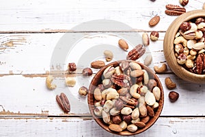 Trail mix with different kinds of nuts in brown wood bowl on scratched white wooden table background, soft daylight. Copy space.