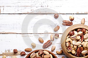 Trail mix with different kinds of nuts in brown wood bowl on scratched white wooden table background, soft daylight. Copy space.