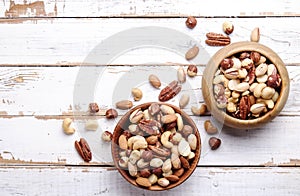 Trail mix with different kinds of nuts in brown wood bowl on scratched white wooden table background, soft daylight. Copy space.