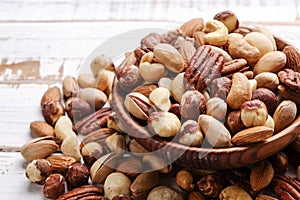 Trail mix with different kinds of nuts in brown wood bowl on scratched white wooden table background, soft daylight. Copy space.