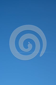 A trail after a plane is seen just under the moon on a clear blue sky