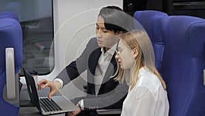 On train an Asian man shows a woman wearing glasses a laptop.