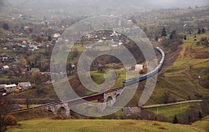 Train Rides On A Winding Railway. Autumn Rural Landscape With Locomotive, Railway Viaduct, Mountains And A Village Located On The