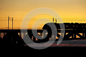 The train riding over the bridge over the river at sunset.