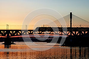 The train riding over the bridge over the river at sunset.