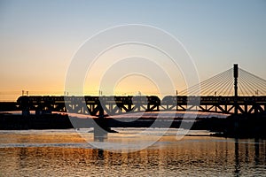 The train riding over the bridge over the river at sunset.