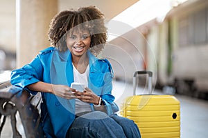 Train Tickets Online. Young Smiling Black Woman Using Smartphone At Railway Station