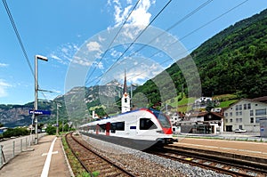 A train travels on the railway curving through a Swiss village with churches on the hillside and majestic Alpine mountains