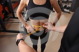 Trainer measuring woman`s waist with tape in gym, closeup photo