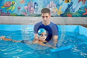 Trainer teaching little boy how to swim in indoor pool with pool floating board