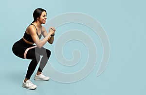 Training for glute muscles. Sporty black woman doing deep squat exercises with elastic bands, blue background