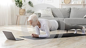 Sporty senior woman doing yoga plank while watching tutorial on laptop