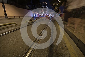 Tram rails on asphalt. People walking and traffic lights on the sidewalk. Filmed at night with long exposure