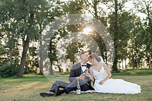 Tranquil, cheerful married couple of bride in wedding dress and groom in suit kissing and drinking bubbly in parkland