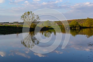 Tranquil landscape at a lake, with the vibrant sky, white clouds and the trees reflected symmetrically in the clean blue water