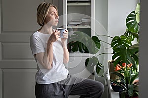 Tranquil middle-aged lady drinks hot drink from mug at windowsill looking out of window