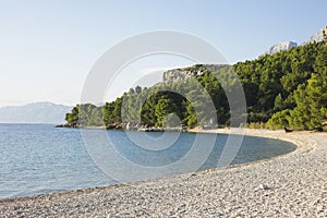 A tranquil pebble beach curves along the clear blue sea in Makarska, Croatia, with lush pines and mountains