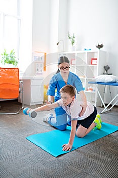 Tranquil short-haired patient holding blue dumbbell while sitting on the mat