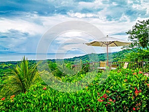 A tranquil terrace with an open umbrella overlooks a lush tropical landscape and the Pacific Ocean, under a dynamic sky