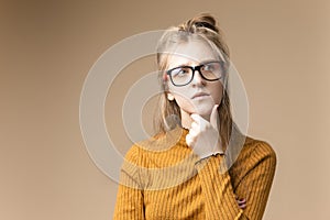 Tranquil Thoughtful Caucasian Blond Female in Yellow Knitted Sweater Posing In Glasses While Touching Chin On Beige Background