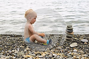 Tranquil toddler meditating on the sea beach. Cute baby boy relaxing and looking to the stone pyramid.