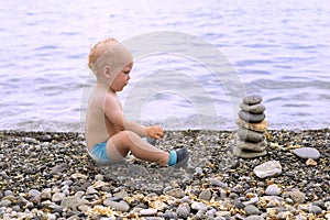 Tranquil toddler meditating on the sea beach. Cute baby boy relaxing and looking to the stone pyramid