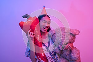 A trans woman wearing a party hat carrying a large teddy bear and chili stuffed toy. Party celebrant scene lit with blue and pink