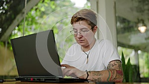 Transgender individual works on laptop in sunny cafe, focused and engaged. Freelancer completes project, exemplifies