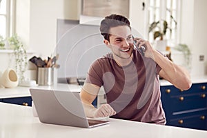 Transgender Man Using Mobile Phone Working From Home Using Laptop On Kitchen Counter