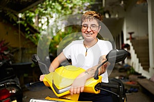 Transgender person smiles, sitting on yellow scooter in tropical location. Short hair, casual style, holding handlebars