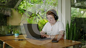 Transgender remote worker types on laptop in plant-filled coworking space. Earphones in, focused, tattooed, embraces