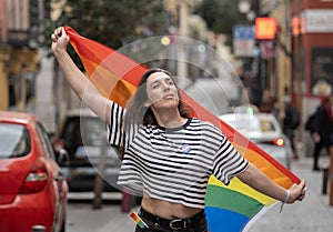 A transgender woman holding a rainbow flag in a city street. The flag is a symbol of the LGBTQ community
