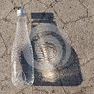 Transparent bottle with drinking water lies on dry ground with cracks casting a wide shadow