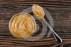 Transparent bowl with peanut butter, spoon with peanut butter on wooden table. Top view