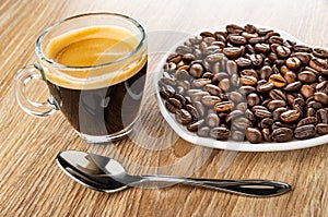 Cup with black coffee, roasted coffee beans in saucer in heart shape, spoon on wooden table