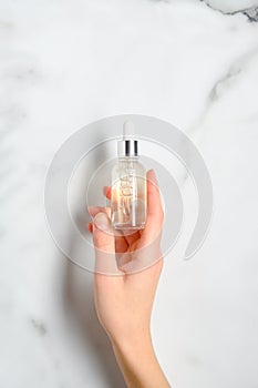 Transparent dropper bottle with serum lotion or essential oil in female hand over marble table. Skin care cosmetics concept