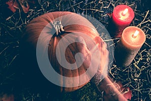 A transparent ghost hand reaching out to touch a big orange pumpkin on dark rustic wood background with hey and candles