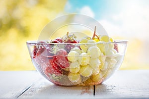 Transparent glass bowl filled with freshly picked strawberries and grapes, washed in water. Blurry bright sky and trees