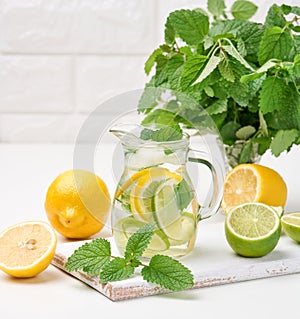 Transparent glass decanter with slices of lemon, lime and mint leaves on a white table, detox. Behind the ingredients for the