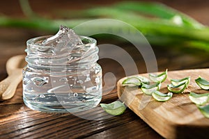 Transparent glass jar of aloe vera gel, sliced aloe leaves on a cutting board. Aloe vera plant on background. Natural remedy