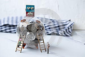 Transparent glass piggy bank, the house is on the top. Miniature people climbing stairs. Money saving ideas for up to buying real