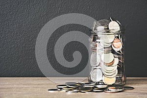 transparent glass savings jar filled with coins on wooden table with black color wall background, copy space